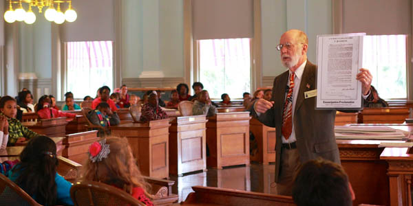 Man in a suit holding a large piece of paper talking to an audience of children